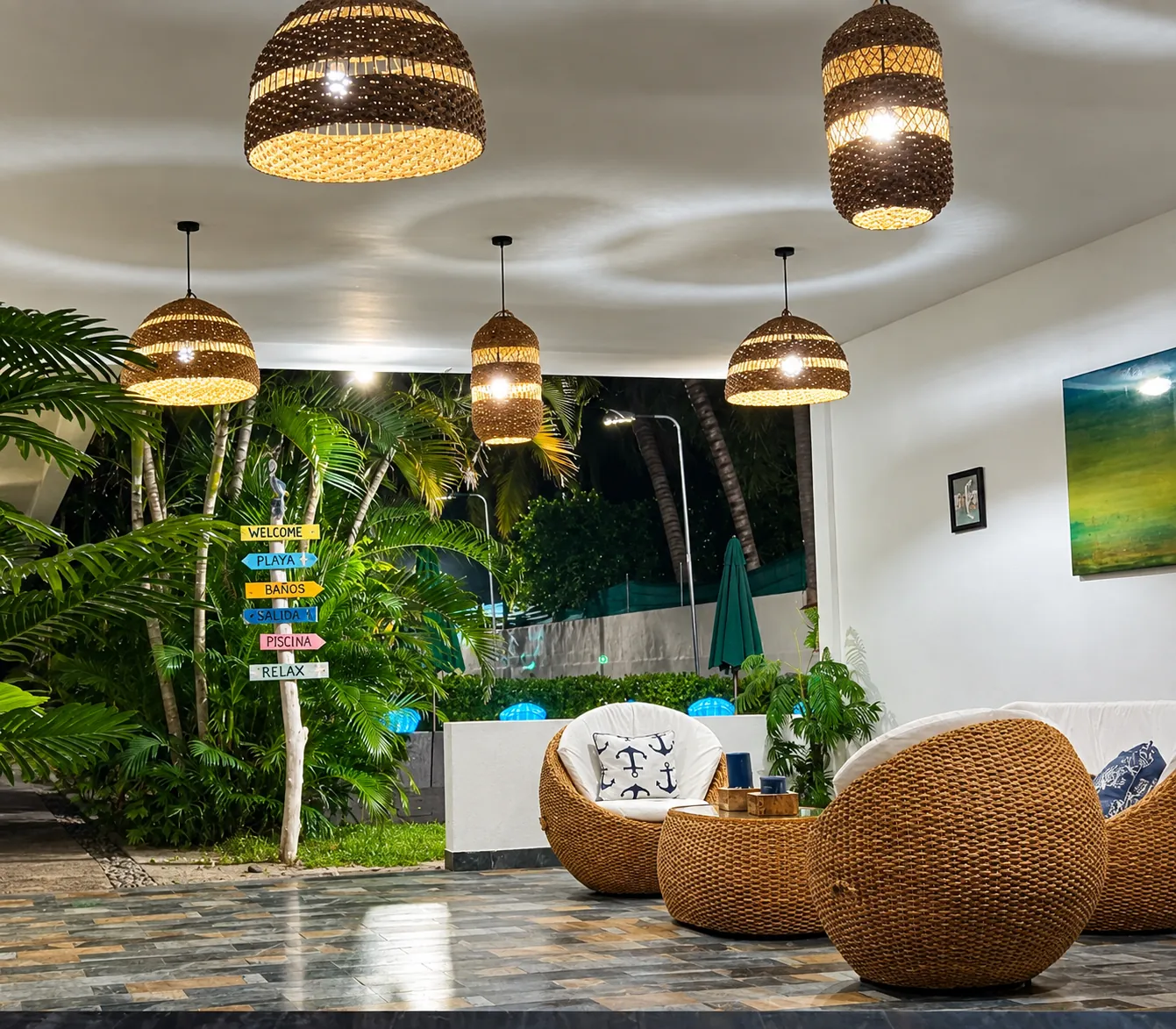 Lounge del hotel de noche con muebles de mimbre y señalización hacia playa, baños y piscina